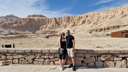 A couple stands before a stone wall with a backdrop of rocky, desert mountains and ancient ruins under the clear blue sky of Thebes. The man dons a black shirt and shorts, while the woman complements her black outfit with a gray hat.