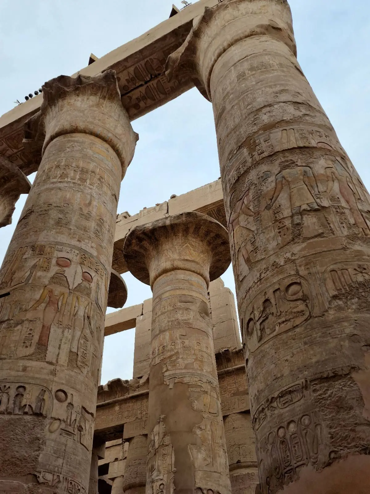 Ancient stone columns towering under a blue sky, adorned with hieroglyphics and carvings. The perspective emphasizes their massive size and intricate details, suggesting an archaeological or historical site.