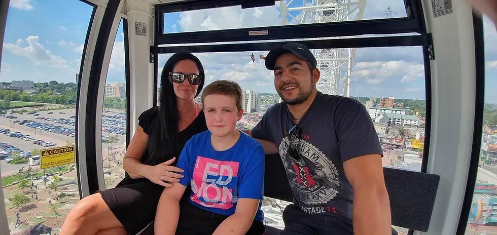 Three people sit inside a Ferris wheel gondola, overlooking a cityscape with blue skies. A "Caution" sign is visible on the window.