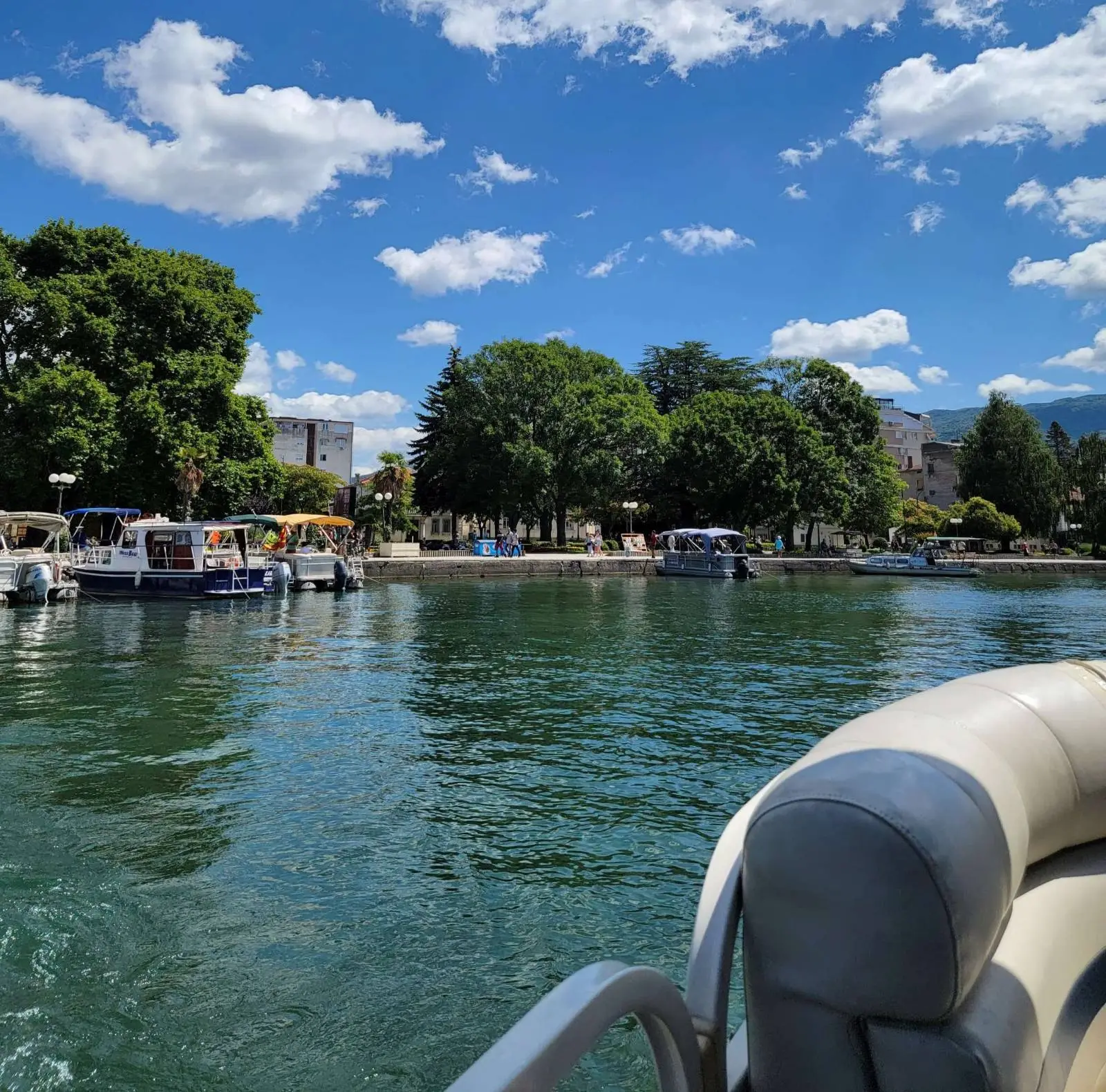 View from a boat on a tranquil lake with clear blue water under a sunny sky with fluffy clouds. Several boats are docked along a tree-lined shore, and a distant town is partially visible through the greenery.