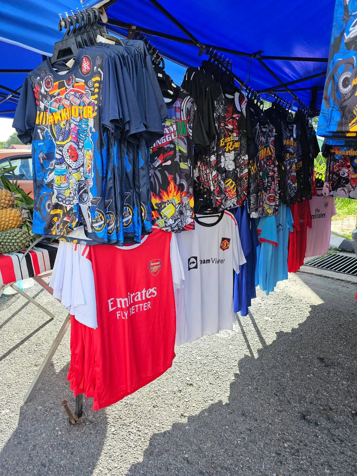A market stall displaying various colorful graphic t-shirts hanging on a rack. The shirts include designs featuring dynamic patterns and motifs. A few shirts have recognizable soccer team logos and colors. The setup is under a blue canopy.