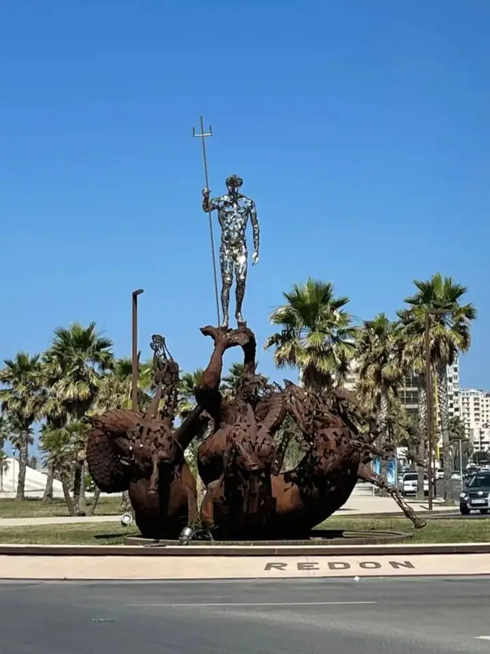 A tall metal statue of a person holding a spear stands atop a large, abstract, rust-colored base in a traffic circle, with palm trees and buildings in the background under a clear blue sky.