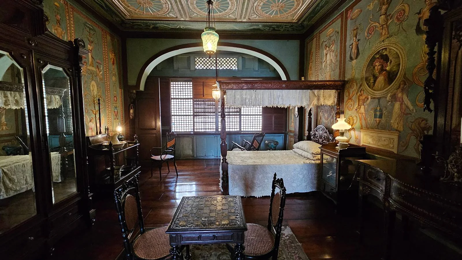 A vintage bedroom with ornate wooden furniture, including a four-poster bed and mirrored wardrobe. The room features intricate wall and ceiling designs, a small table with chairs, and soft lighting from lamps and a hanging fixture.