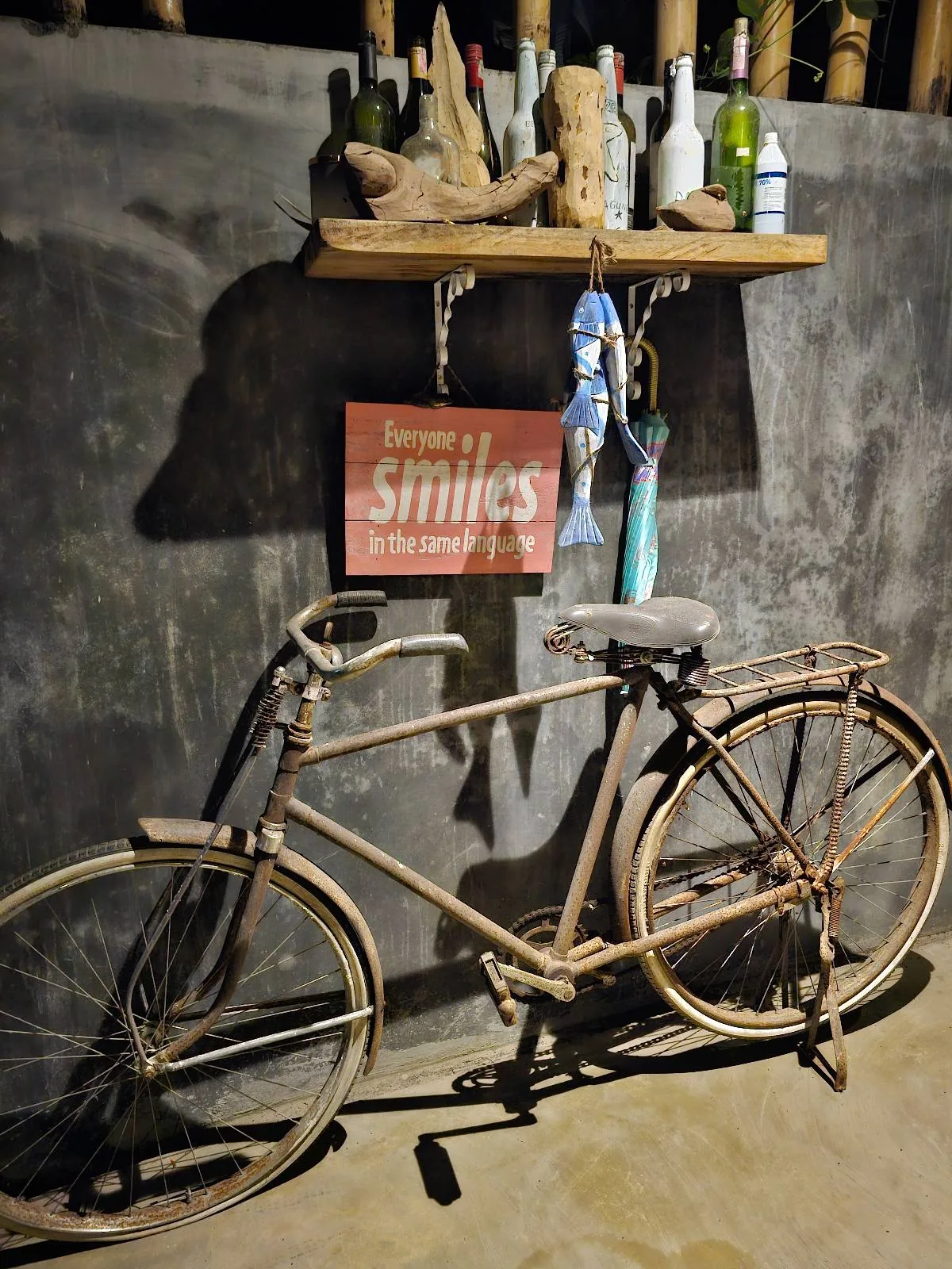 A vintage bicycle is leaning against a dark wall. Above it, a wooden shelf holds various bottles and figurines. A sign with the word "Smiles" hangs on the wall. The scene is lit by warm lighting, creating a cozy ambiance.