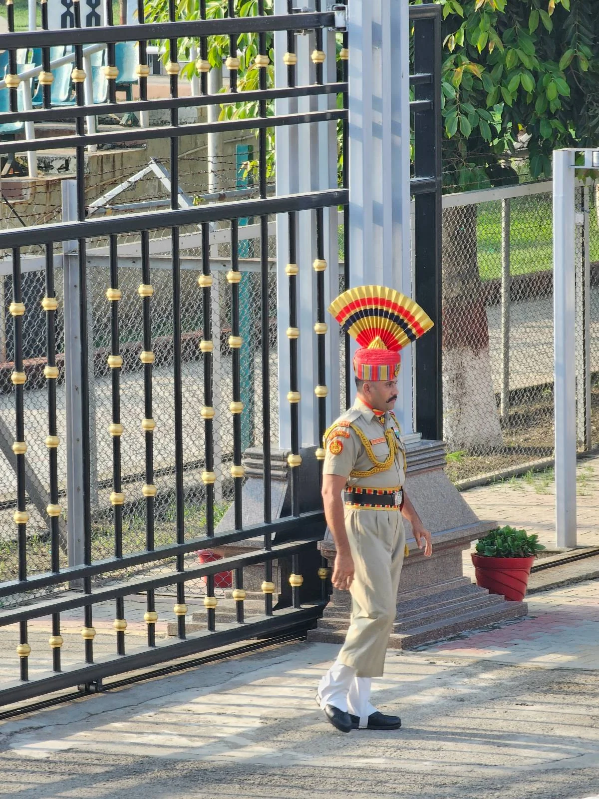 A uniformed border guard wearing a colorful, fan-shaped headdress stands near a black iron gate with gold accents and a red potted plant nearby on a sunny day.