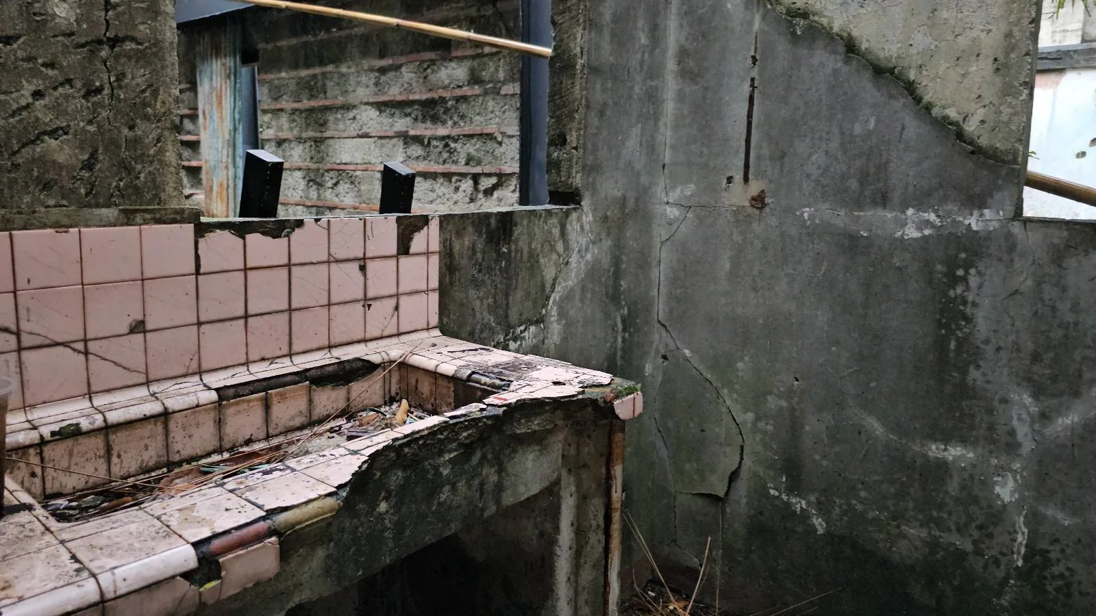 A dilapidated structure with a broken sink and pink tiles. The walls are cracked and weathered, and debris is scattered on the floor. Wooden beams lean against the walls, adding to the abandoned and deteriorating atmosphere.