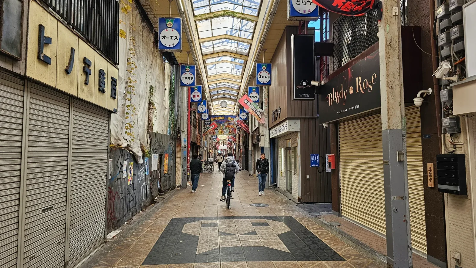 A covered shopping street in Japan with closed shop shutters, a few people walking, and a cyclist riding down the center. Colorful signs and banners hang from the ceiling, and natural light enters through the glass roof.