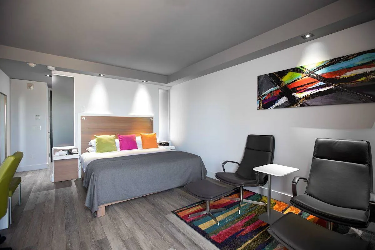 Modern hotel room with a large bed featuring colorful pillows. Two sleek black chairs and a small white table are arranged on a vibrant rug. An abstract painting decorates the wall, and the room is lit with soft, recessed lighting.