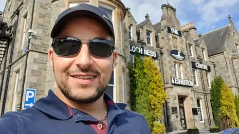 Leon standing near the Loch Ness visitor centre with the loch and stone buildings behind him.