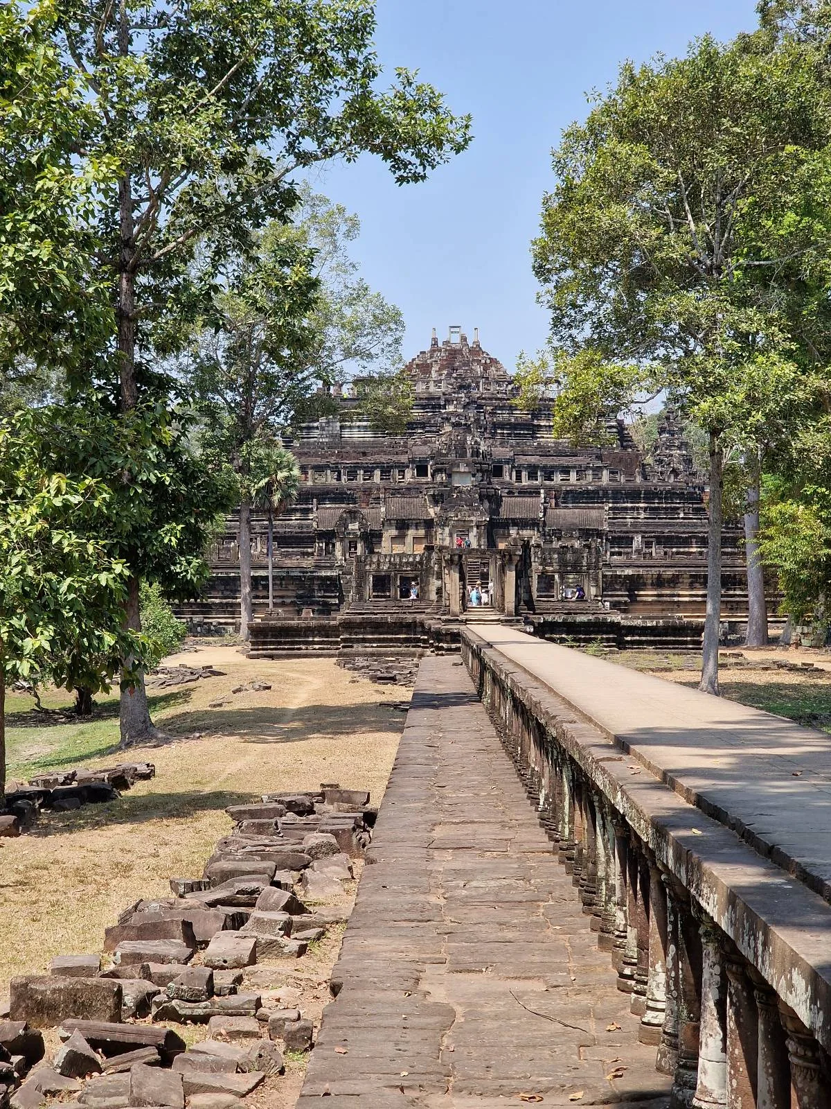 Ancient stone temple with detailed carvings, surrounded by green trees under a clear blue sky, viewed from a stone walkway leading to its entrance.