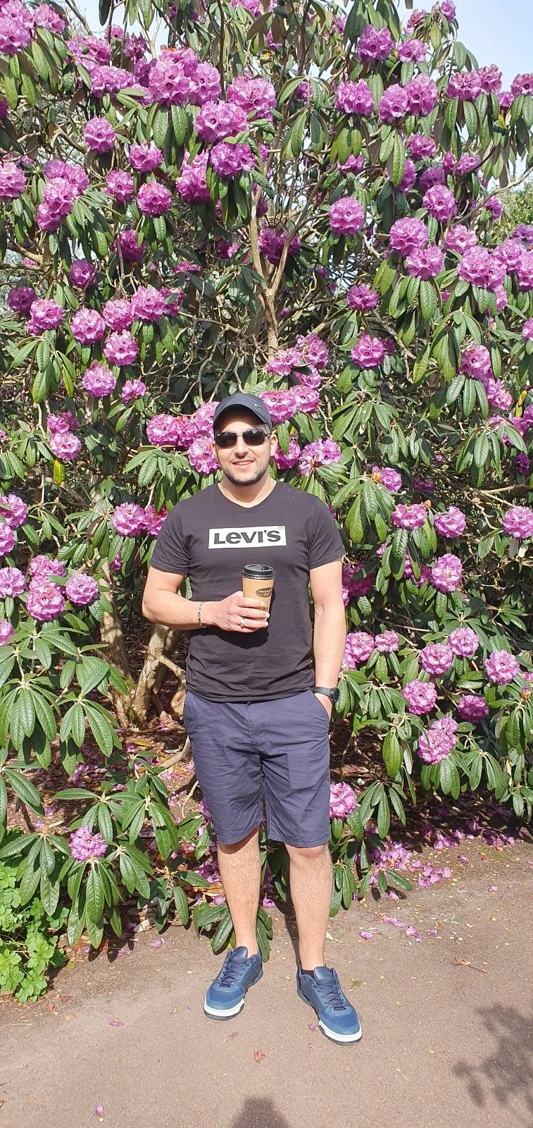 Man standing in front of a large flowering bush with pink blossoms, wearing a cap, T-shirt, shorts, and sneakers, holding a drink.