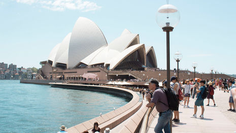 People walk along a waterfront promenade near the Sydney Opera House on a sunny day, with the iconic white sails of the building visible and clear blue water to the left.
