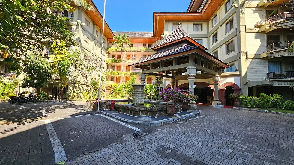 Courtyard of a tropical hotel with a central fountain, surrounded by lush greenery and a multi-story building under a clear blue sky.