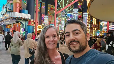 A smiling couple takes a selfie in a colorful, bustling indoor amusement park with a brightly lit Ferris wheel and crowds of people in the background, capturing memories while enjoying great food options and theme park ticket value.
