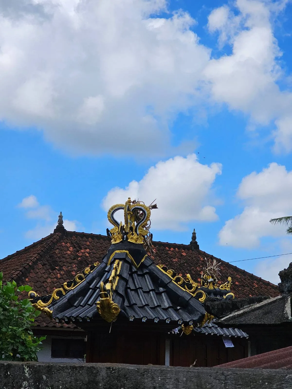 A traditional building with ornate, gold-accented black roof tiles under a blue sky with white clouds. The roof has intricate, decorative elements at the edges and a lush green tree in the foreground.