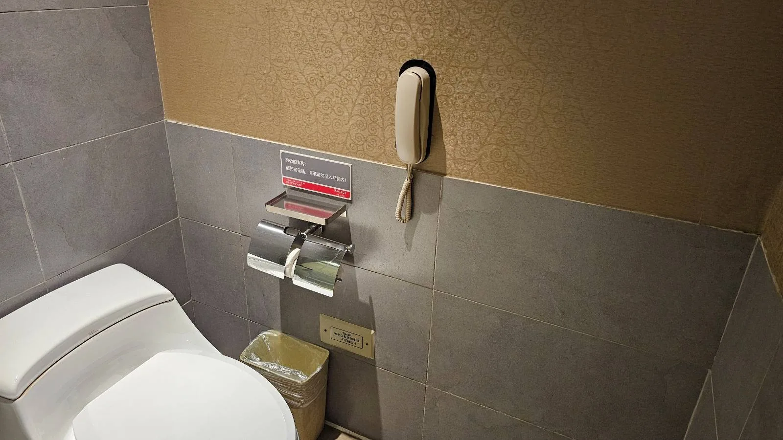A bathroom with a toilet, two rolls of toilet paper, a wall-mounted telephone, a small trash bin, and a sign above the toilet paper dispenser.