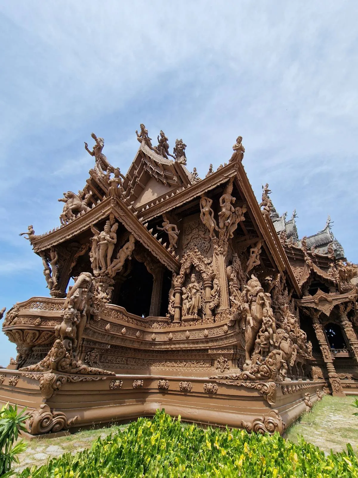 A large, intricately carved wooden temple with ornate details and multiple pointed rooftops stands against a blue sky, surrounded by green plants at its base.