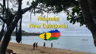Cloudy beach scene in Nouméa, New Caledonia, with people walking by the shore and large text over a bay framed by trees.