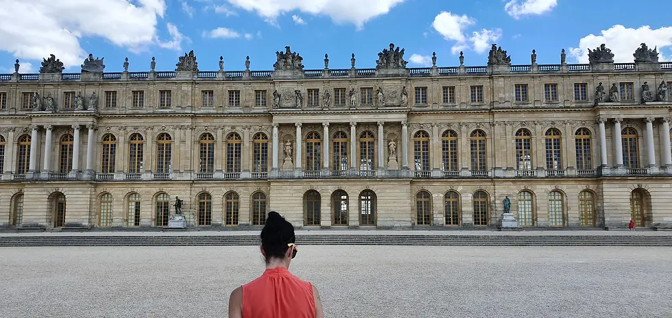 Woman in red top stands facing the Palace of Versailles, featuring statues and ornate architecture under a blue sky with clouds.