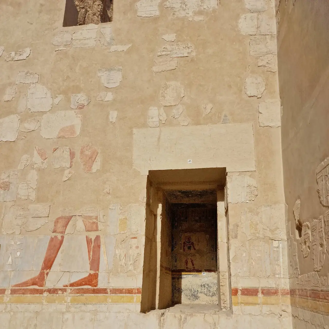 An ancient stone wall with faded carvings and paintings, featuring a rectangular doorway leading to a small alcove. Inside, there is a partially visible relief. The wall shows signs of age and weathering.