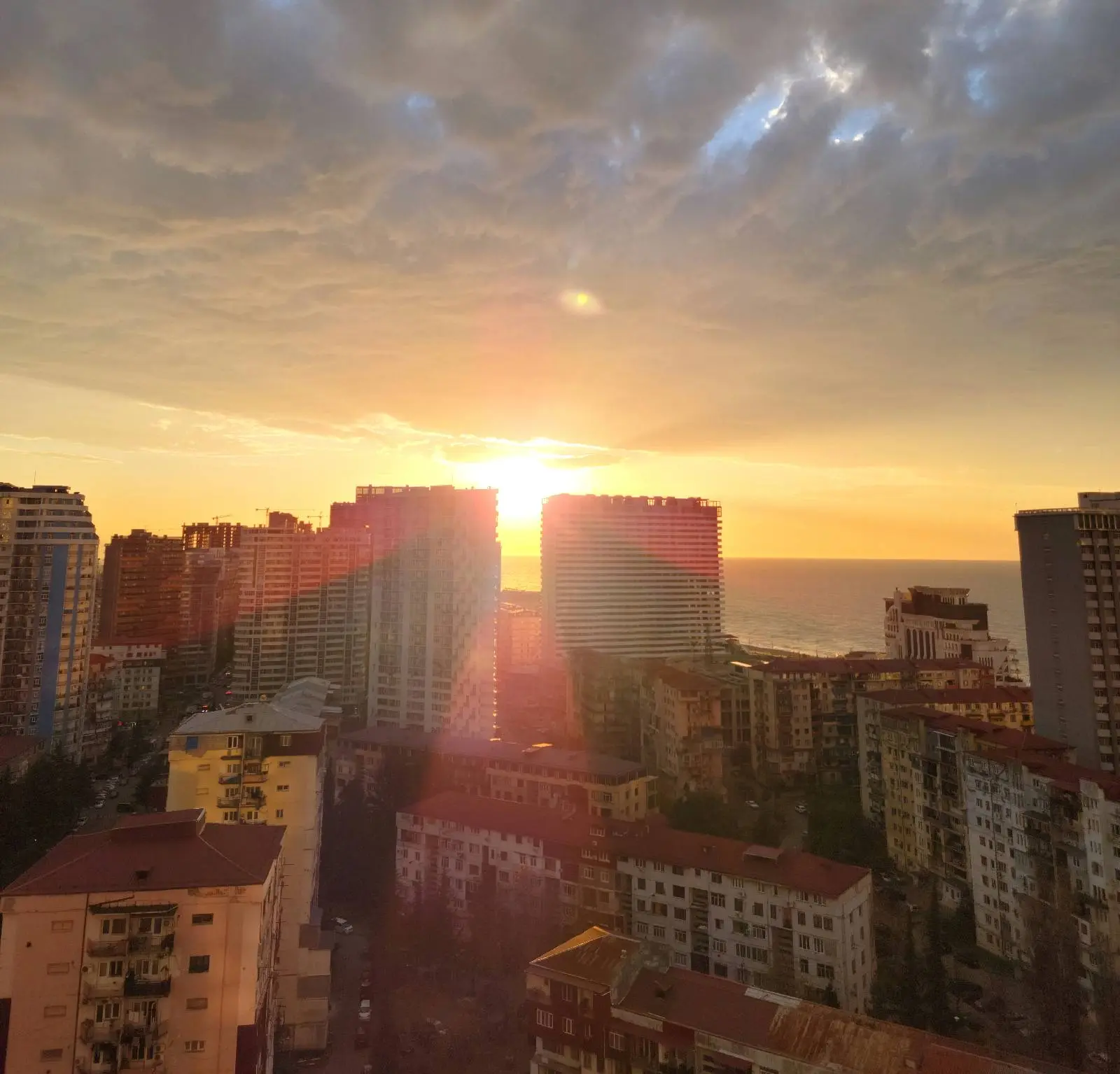 Sunset over a cityscape with tall buildings and red rooftops, sunlight streaming through gaps between the buildings, and a partly cloudy sky above.