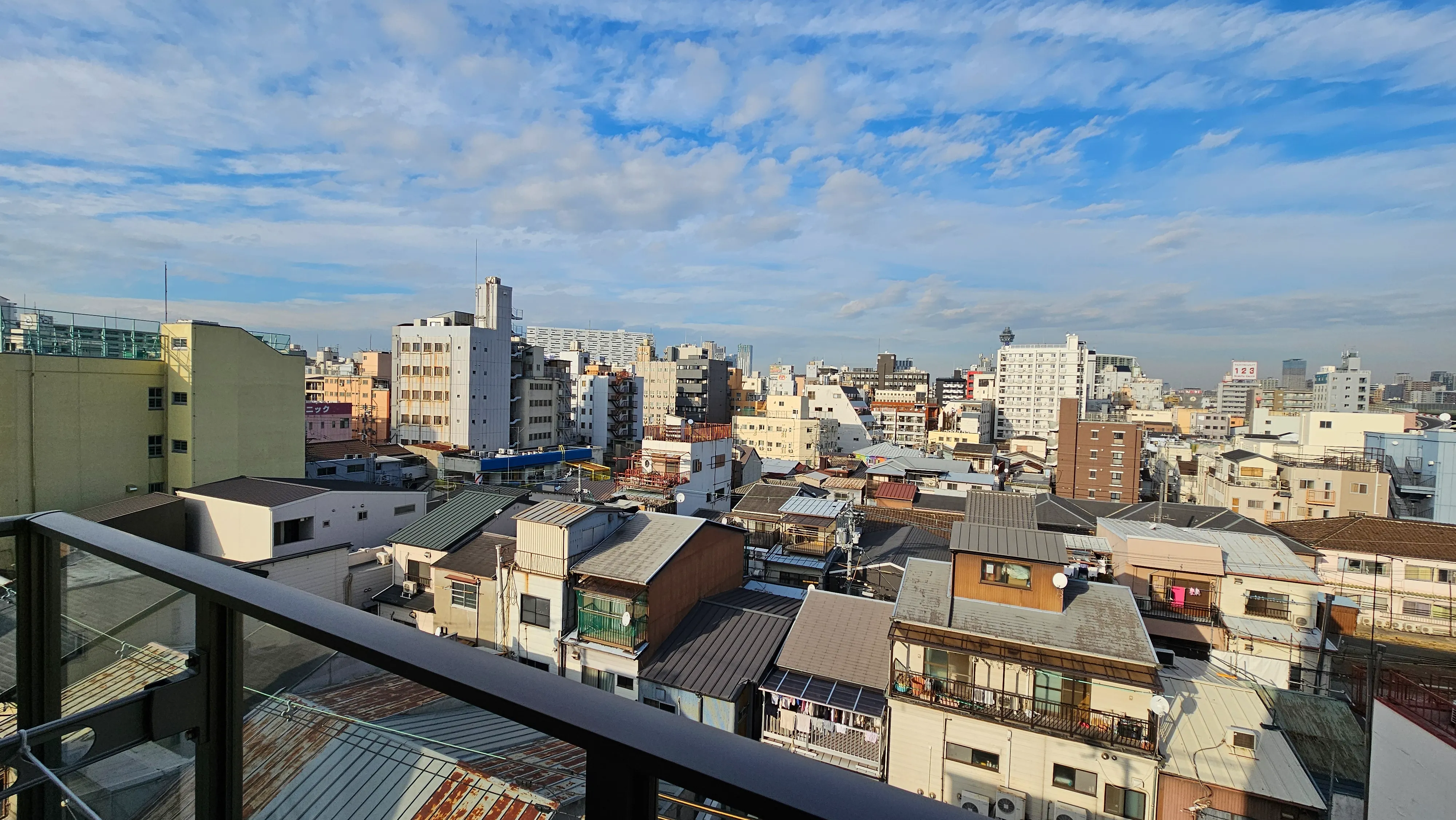 Cityscape view from a balcony showing a mix of low-rise and mid-rise buildings under a blue sky with scattered clouds. The image captures an urban residential area with various rooftops.