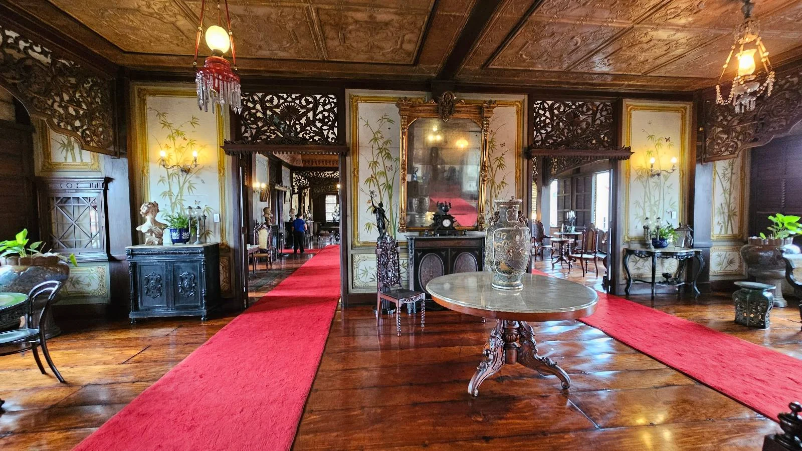 Opulent interior with decorative woodwork, ornate chandeliers, and red carpets on polished wooden floors. A central round table is framed by elegant furnishings and intricate wall designs, creating a luxurious atmosphere.