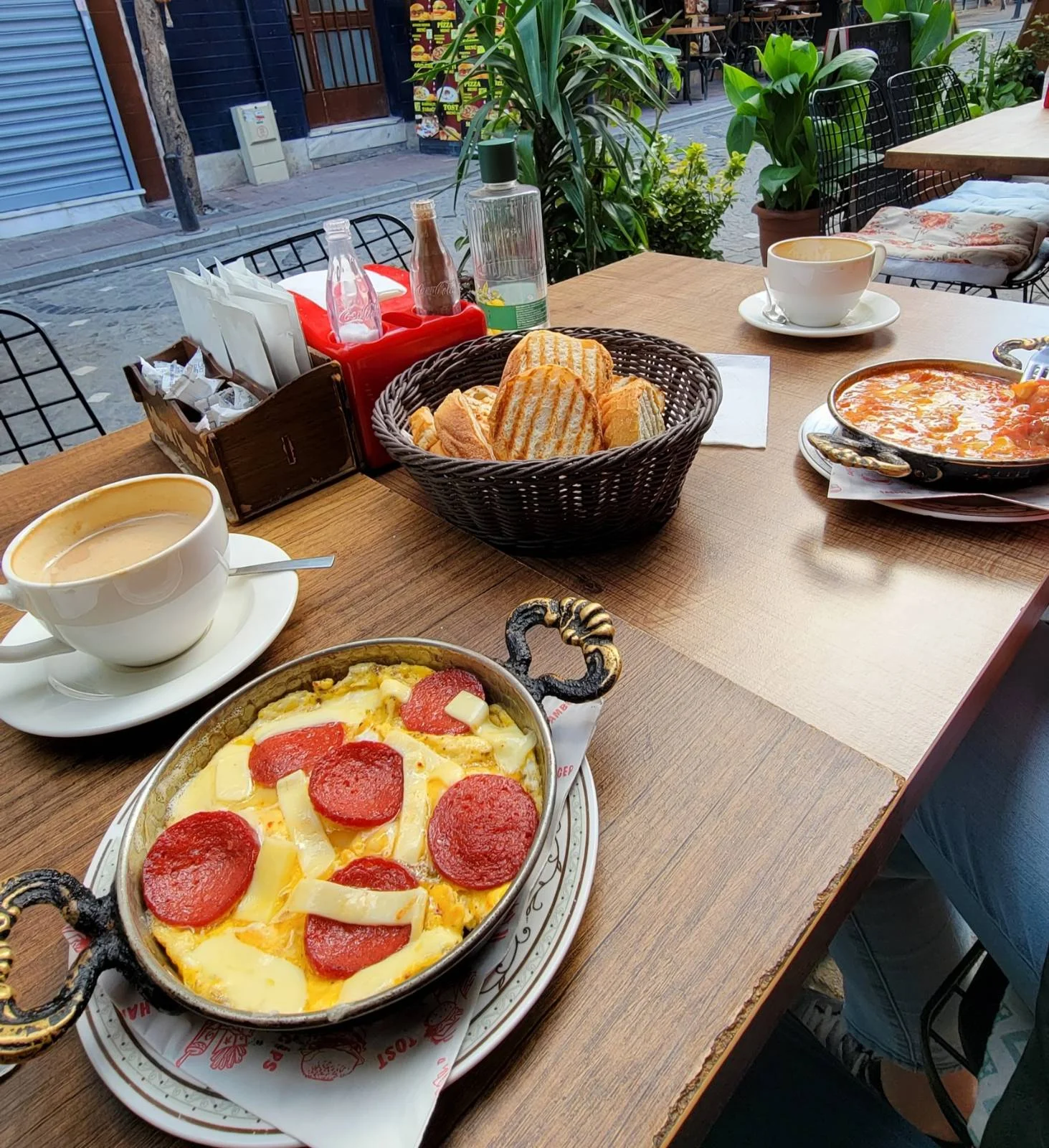 A platter with cheese, salami, cucumbers, olives, tomatoes, and jam. A bowl of bread and a drink are in the background.