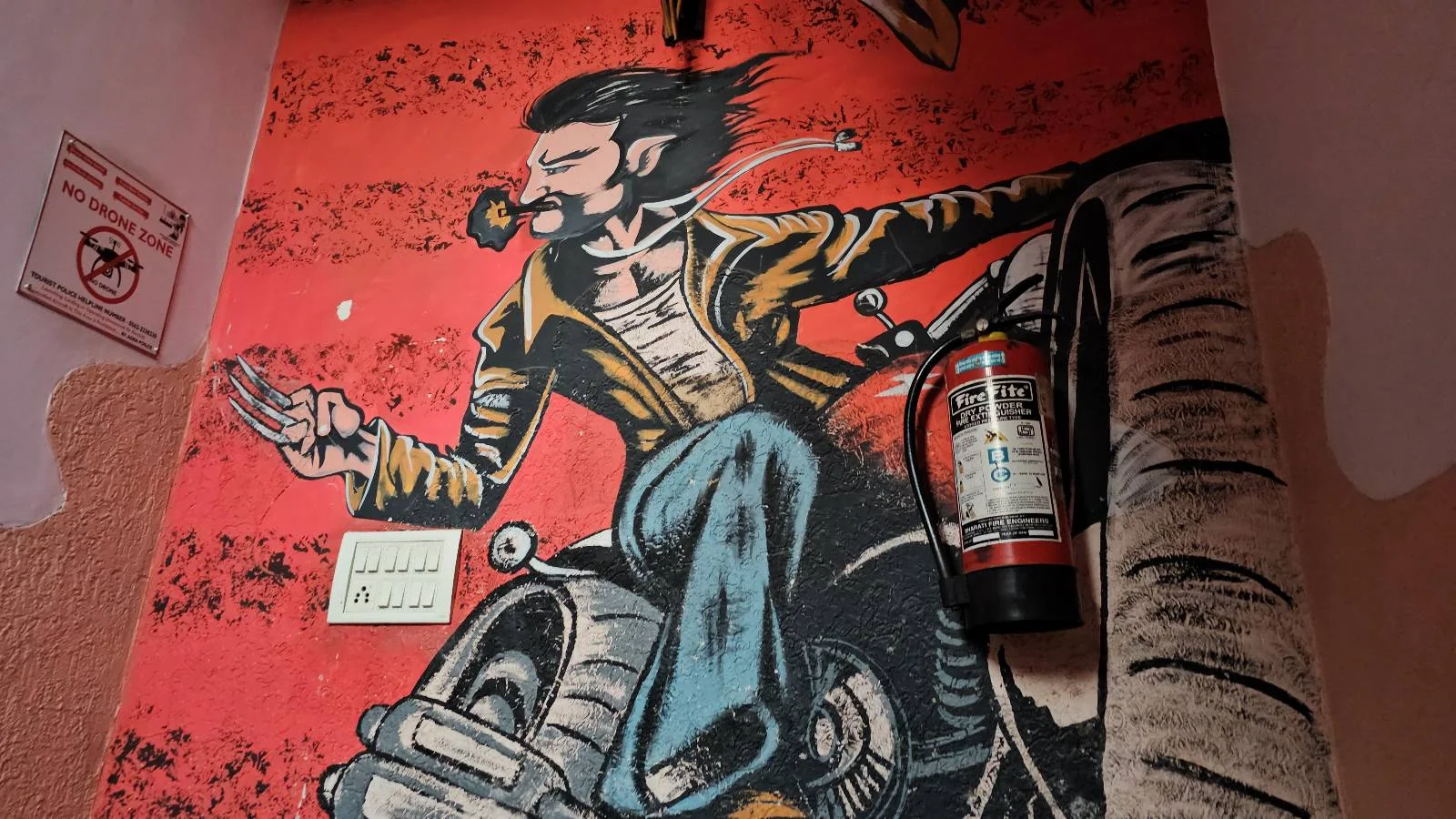 A mural of a man with facial hair riding a motorcycle, wearing a yellow jacket and blue pants. He's holding up three fingers on one hand. A fire extinguisher is mounted on the wall next to the mural.