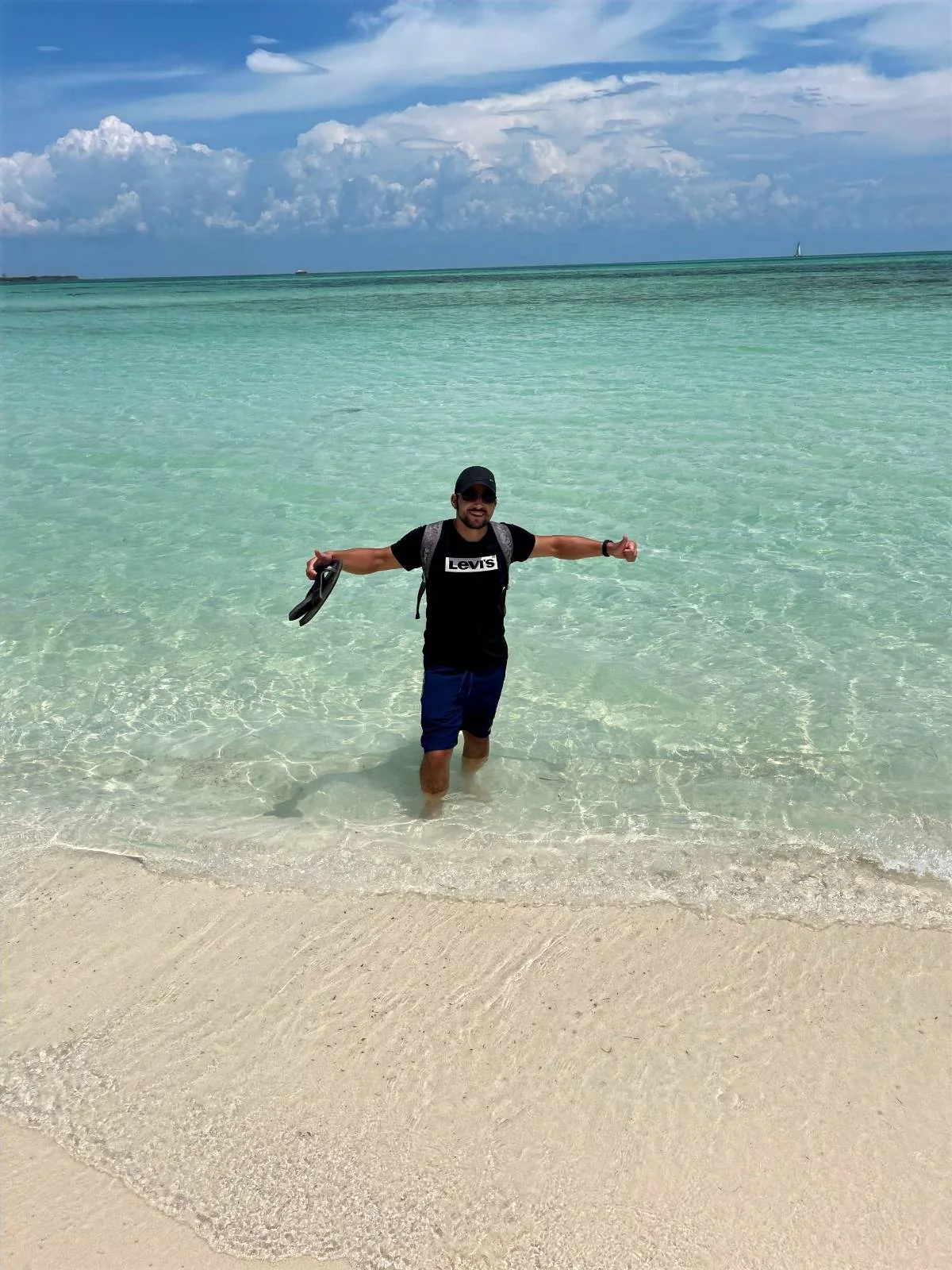 A person stands in shallow, clear turquoise ocean water near a sandy beach, holding shoes in one hand. They wear a black cap, sunglasses, a black T-shirt, and blue shorts. The sky is partly cloudy with a distant horizon.