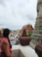 Woman in a pink top and black cap looks out over ornate temple roofs and a tall, intricate pagoda. Cloudy sky adds a serene mood.