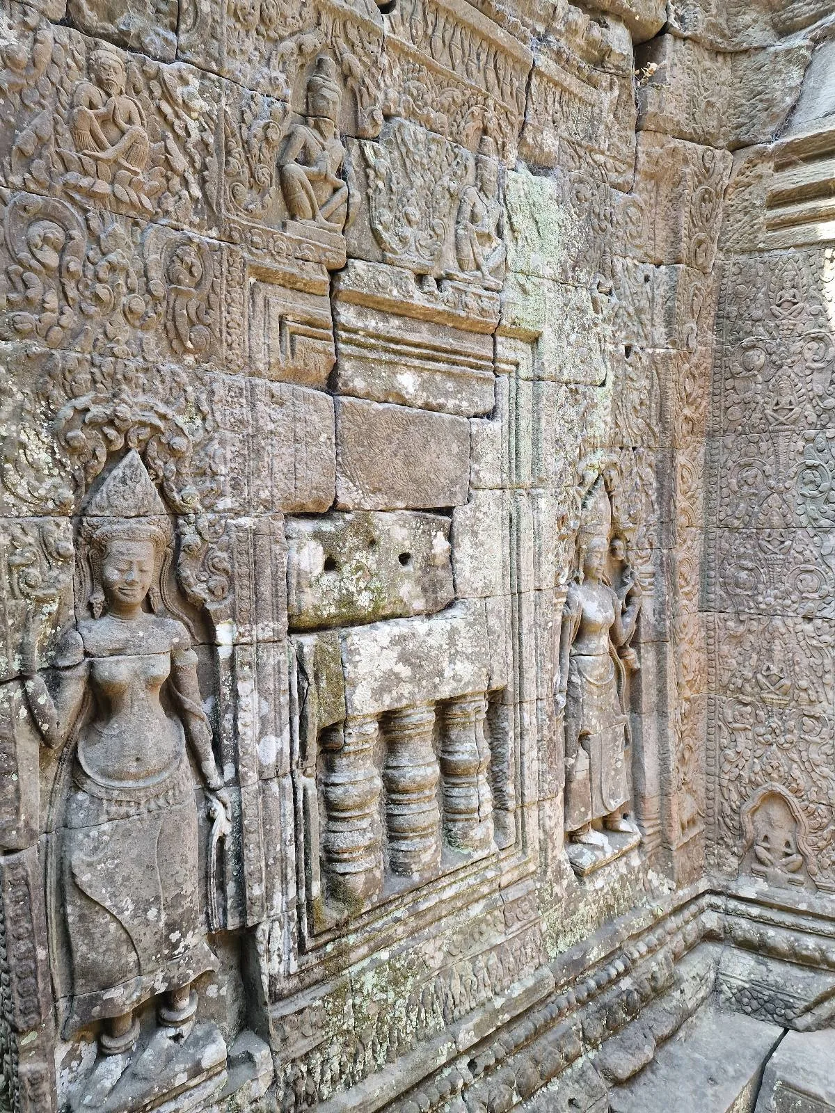 Stone wall carvings at an ancient temple depict detailed human figures and intricate patterns, showing weathered but elaborate artwork on the surface. The carvings appear to be part of historic architecture.