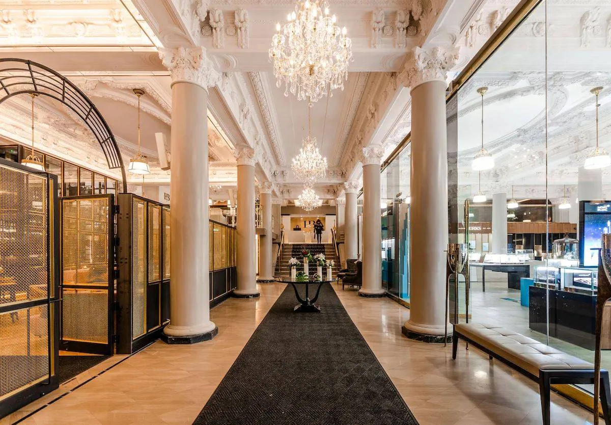Elegant hotel lobby with chandeliers, marble columns, black carpet, and modern seating. Glass displays on right, person at stairs. Luxurious ambiance.