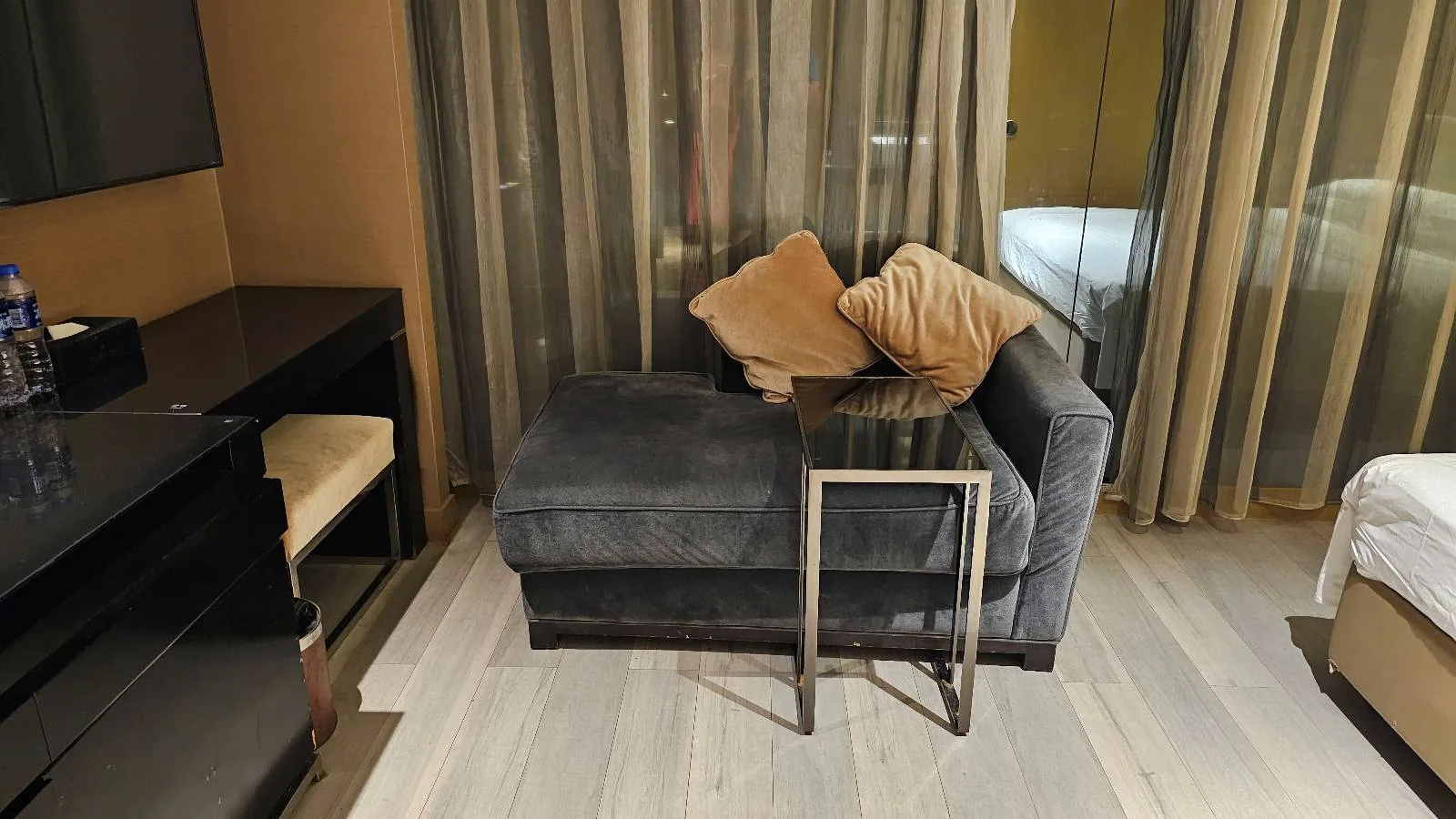 A modern hotel room features a dark grey chaise lounge with light brown pillows, a small metal side table, a desk with a chair, and a bed partially visible on the right, all on light wood flooring.