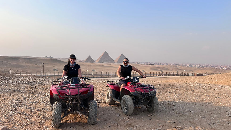 Two people on red ATVs in a desert with the Pyramids of Giza in the background. Clear sky and rocky terrain.