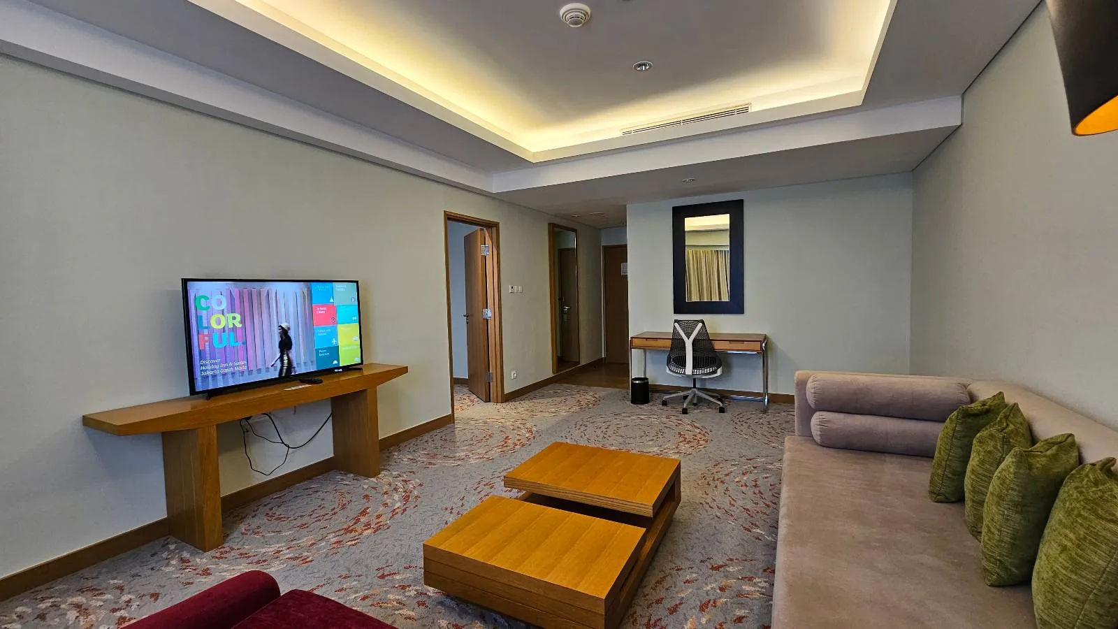 Modern living room with a wall-mounted TV, beige sofa with green pillows, two wooden coffee tables, a desk with a chair, a wall mirror, and recessed ceiling lighting. The floor is carpeted with a patterned design.