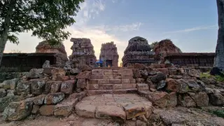 Ancient stone ruins of Phnom Kraom with a stepped pathway leading to large, intricate structures under a blue sky with soft clouds. Trees flank the sides, and the sun is setting in the background, creating a warm, serene atmosphere reminiscent of the era of Khmer King Yasovarman I.