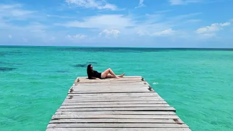 A person in a black swimsuit lies on a wooden pier extending into the clear, turquoise waters off Passion Island, Mexico. Beneath the bright blue sky with fluffy white clouds, this serene scene promises a relaxing day in paradise.