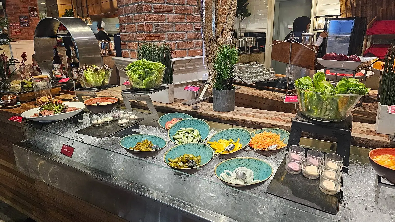 A buffet setup featuring a variety of fresh salad ingredients in turquoise bowls, including sliced cucumbers, bell peppers, and olives, with leafy greens displayed in baskets. The arrangement is on a dark countertop, accented with small potted plants and a brick wall in the background.