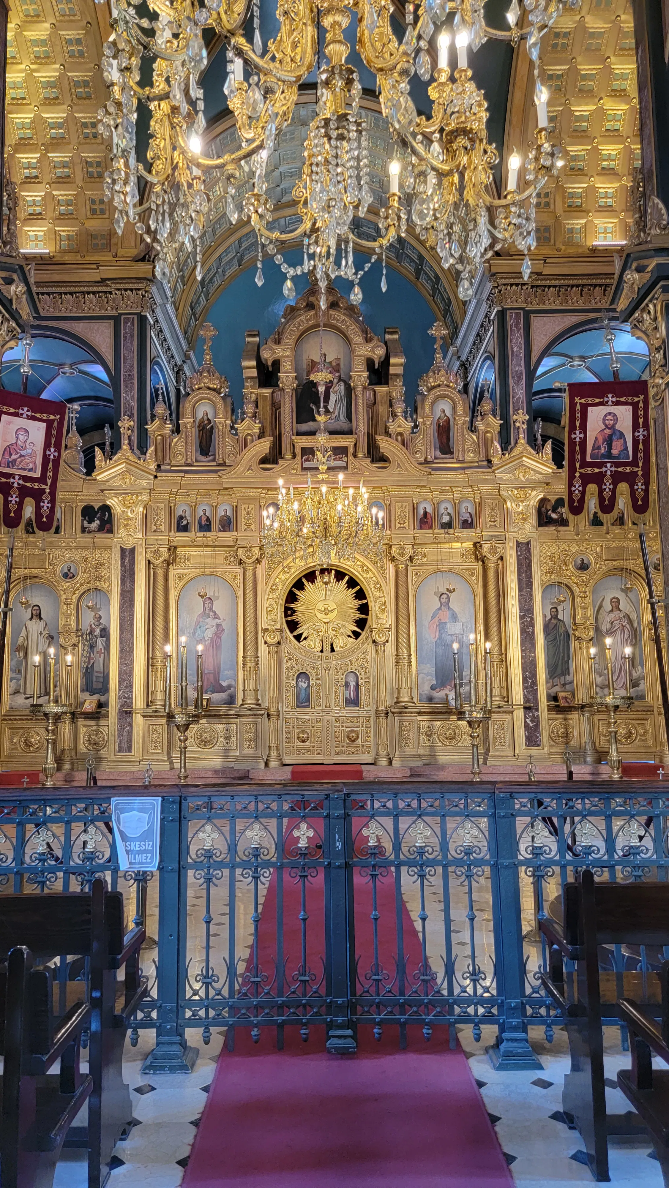 An ornate church altar adorned with gold decorations, religious icons, and paintings, framed by a decorative gate and red carpet. Chandeliers hang above, and rows of pews are visible on either side.