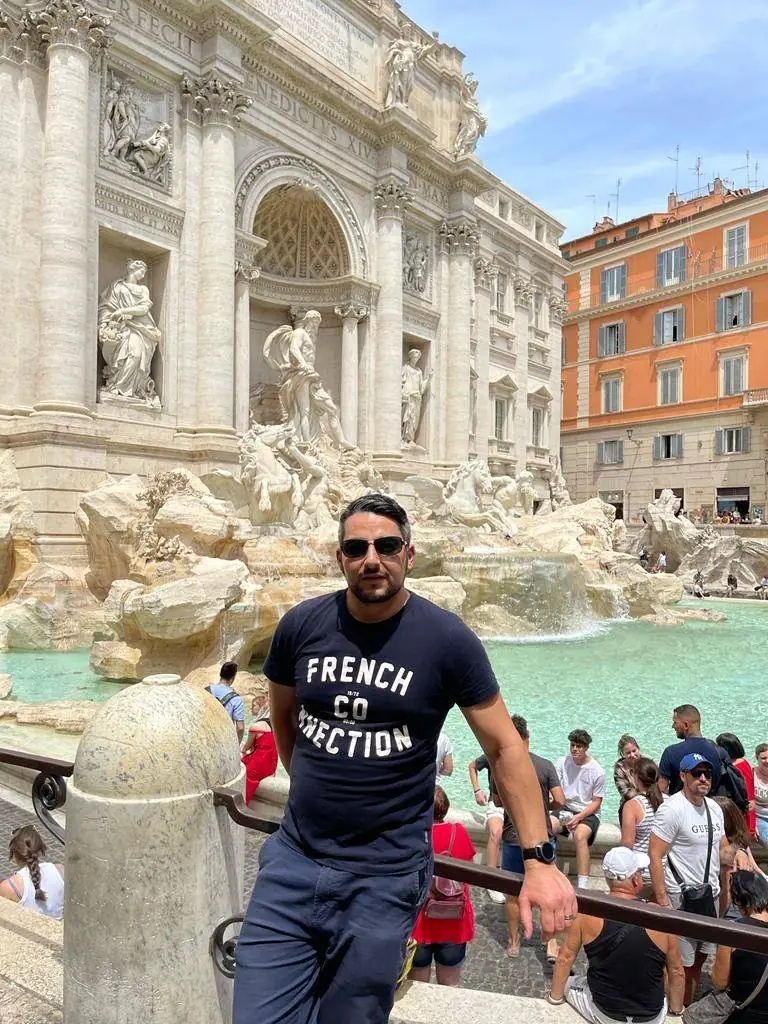 A man wearing sunglasses and a navy T-shirt stands in front of the Trevi Fountain in Rome, Italy, with a crowd of tourists and historic buildings visible in the background.