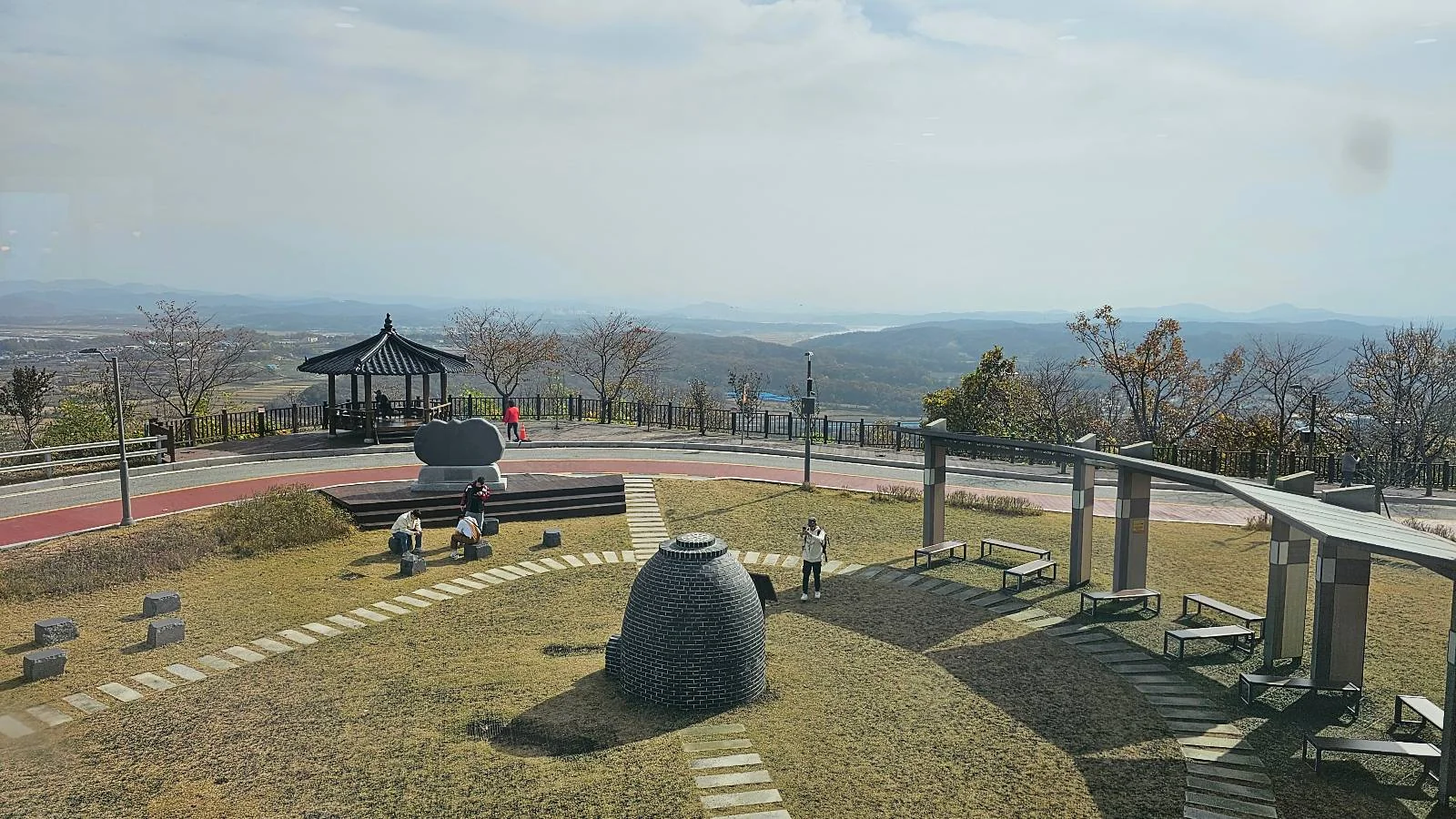 A scenic viewpoint with distant hills under a cloudy sky. In the foreground are stone sculptures and a path, leading to a circular pavilion. Sparse trees dot the landscape, with a vast horizon stretching beyond.