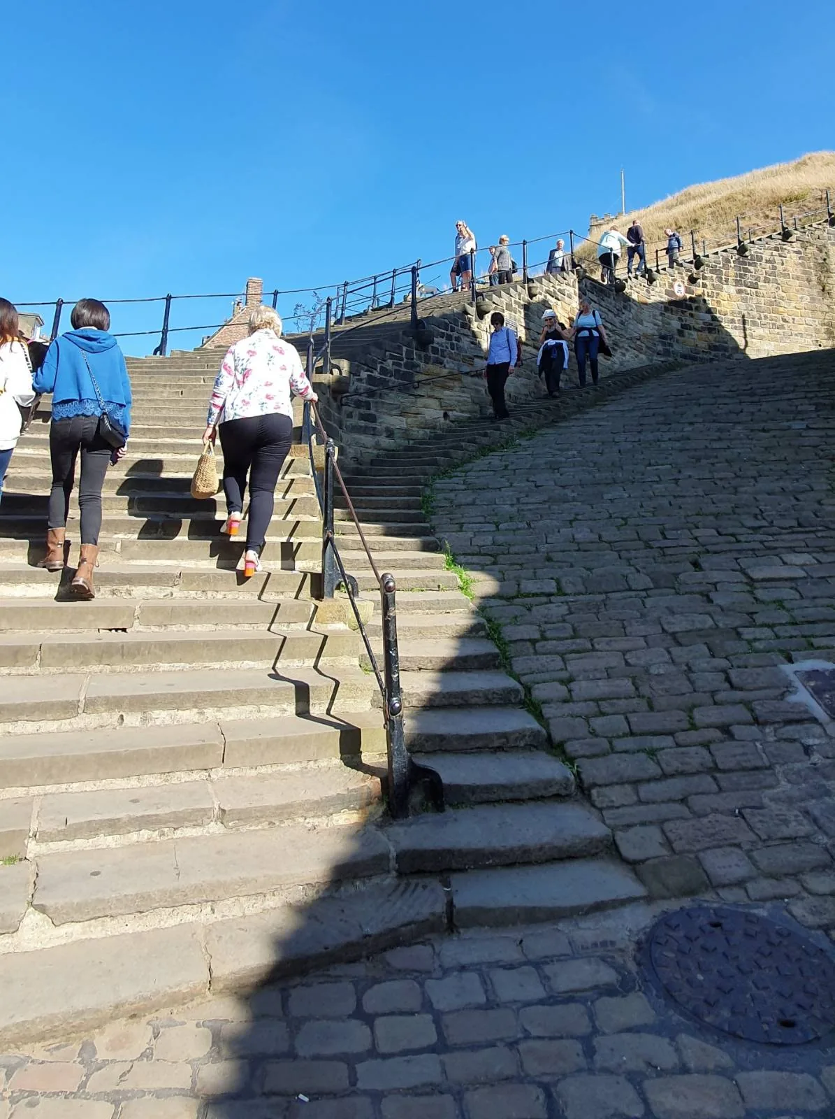 People walking up a wide stone staircase on a sunny day. The sky is clear and blue. A metal handrail runs alongside the stairs, and more people can be seen ascending further up in the distance.