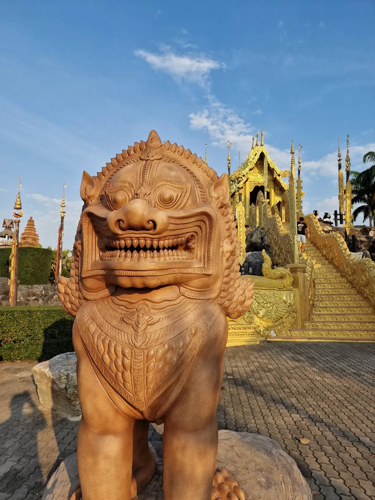 Stone statue of a mythical lion with an ornate temple entrance and golden staircase in the background, set against a clear, blue sky.
