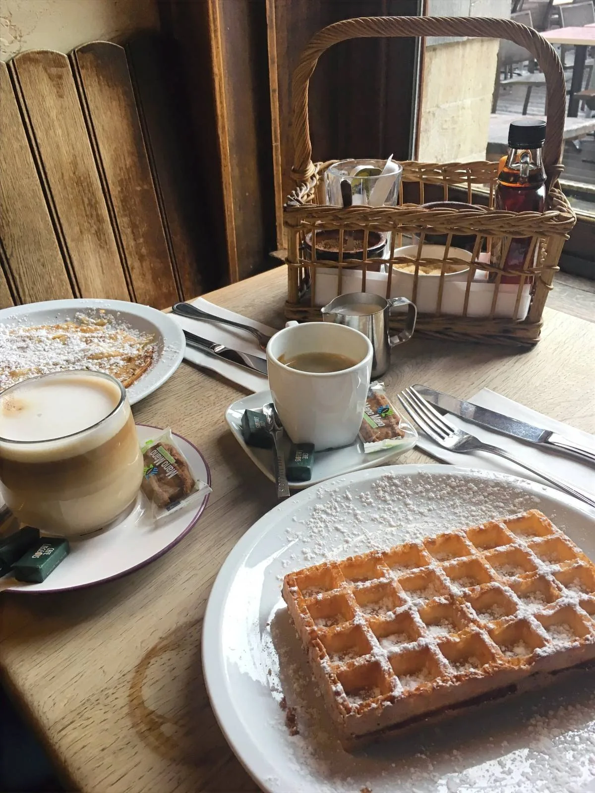 A table set with a plate of powdered sugar-covered waffles and a cup of coffee. Another plate holds a cappuccino with a biscuit. Cutlery is laid neatly, and a small basket holds condiments. Sunlight streams through a window onto the wooden table.
