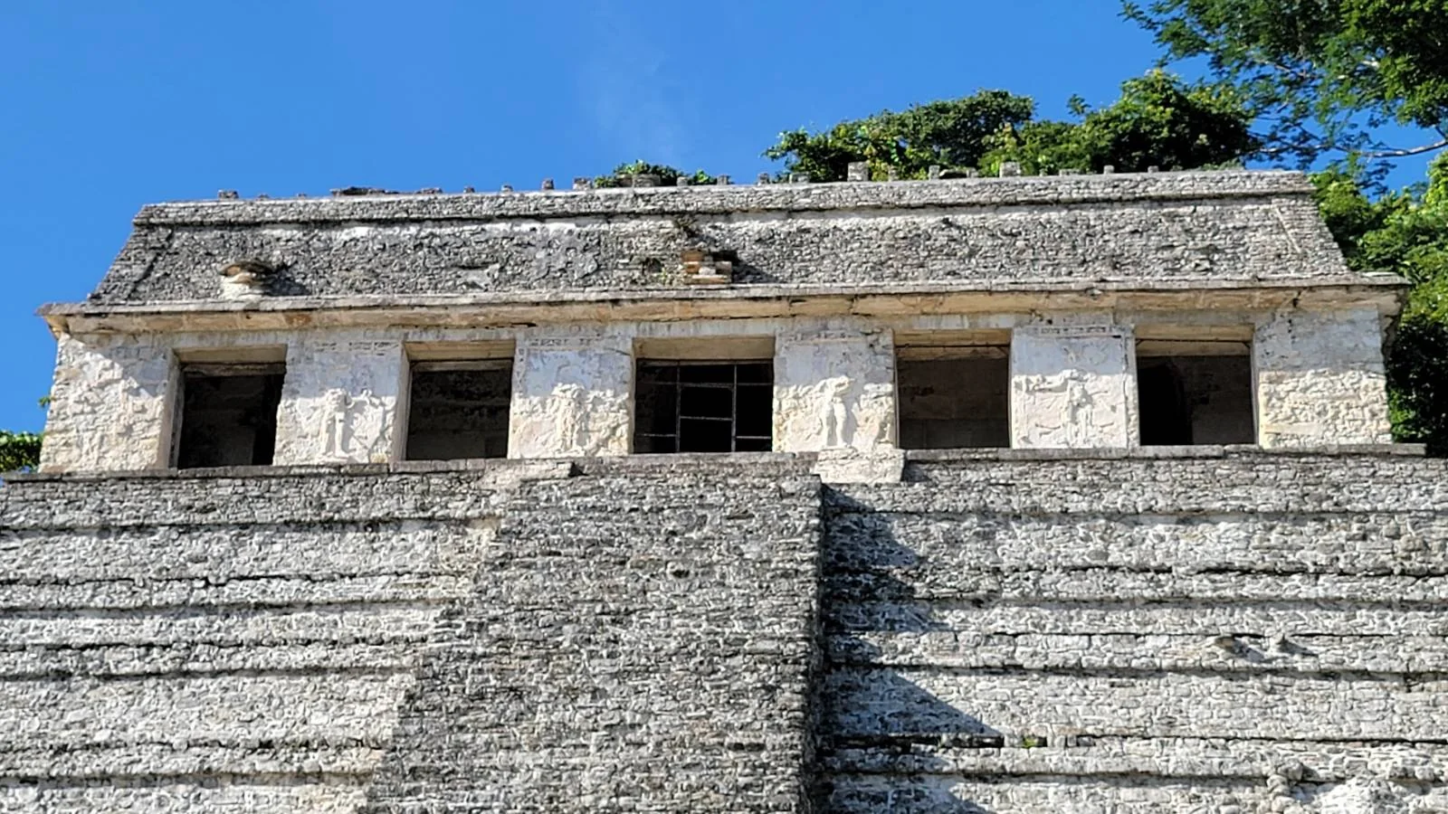 Ancient stone pyramid with a staircase leading to a structure featuring multiple rectangular openings. The structure is set against a backdrop of clear blue sky and lush green foliage at the top.