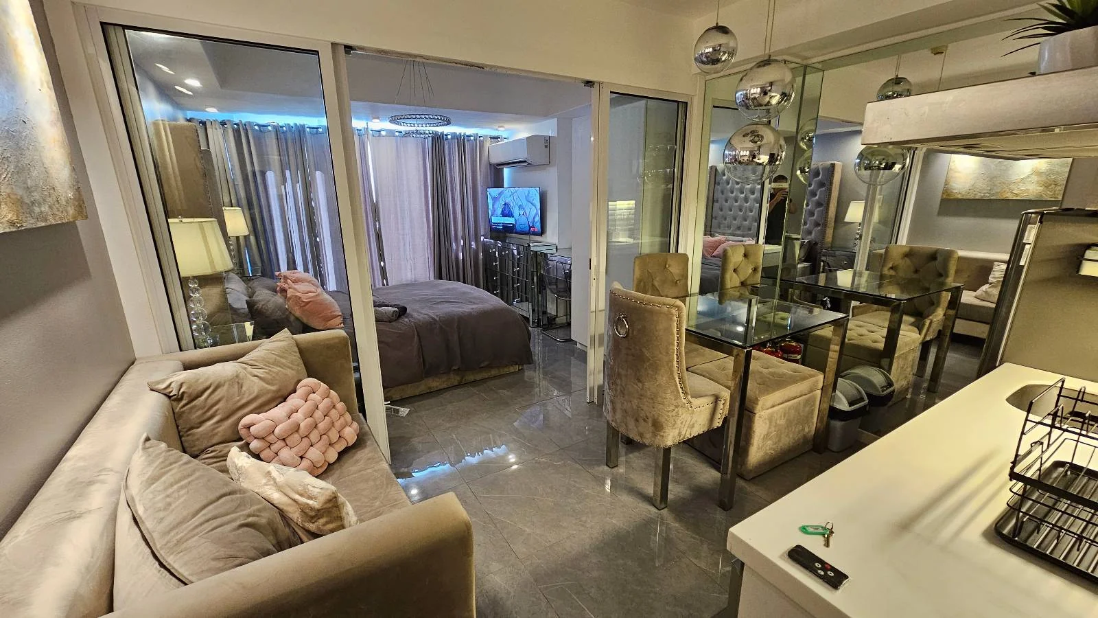 Modern living room and bedroom with glass divider. Beige sofa, plush pillows, dining table, TV on, and mirrored wall. Cozy ambiance.