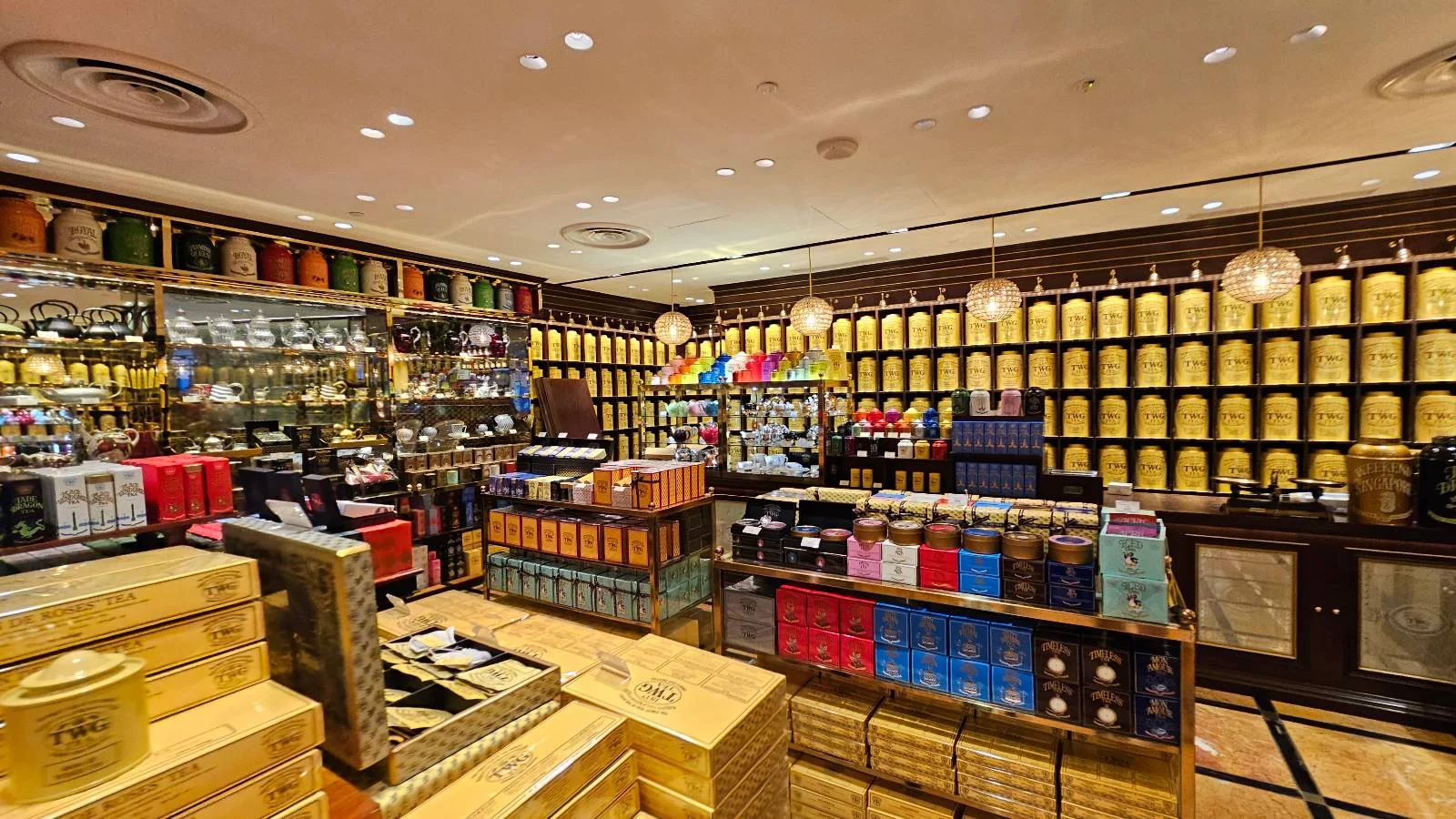 A brightly lit tea shop with shelves full of tea canisters lining the walls. Various tea boxes and tins are displayed on wooden tables. The warm, inviting atmosphere includes pendant lights hanging from the ceiling.