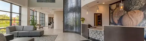 A modern, spacious lobby in luxury resorts with a high ceiling features a water wall, contemporary seating, and a reception desk with pendant lights. Large windows on the left allow natural light to illuminate the area, creating a welcoming atmosphere.