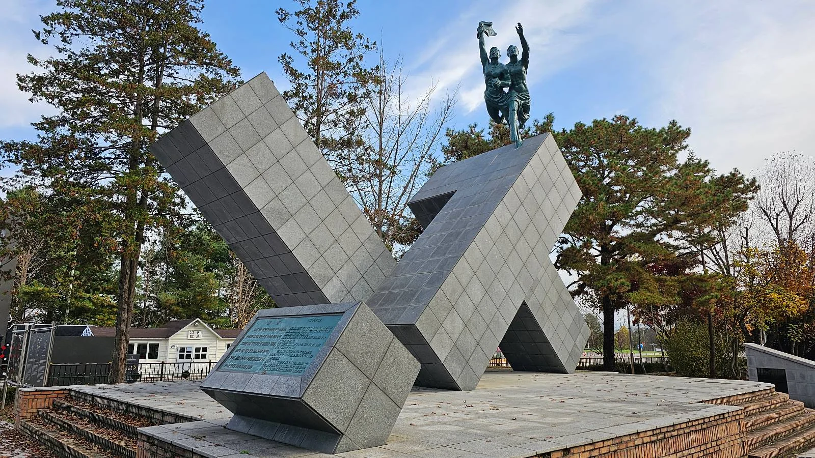 A large geometric monument with intersecting angular blocks stands in an outdoor area. Atop the structure is a dynamic statue of a human figure reaching upward. Trees and a building are visible in the background under a partly cloudy sky.